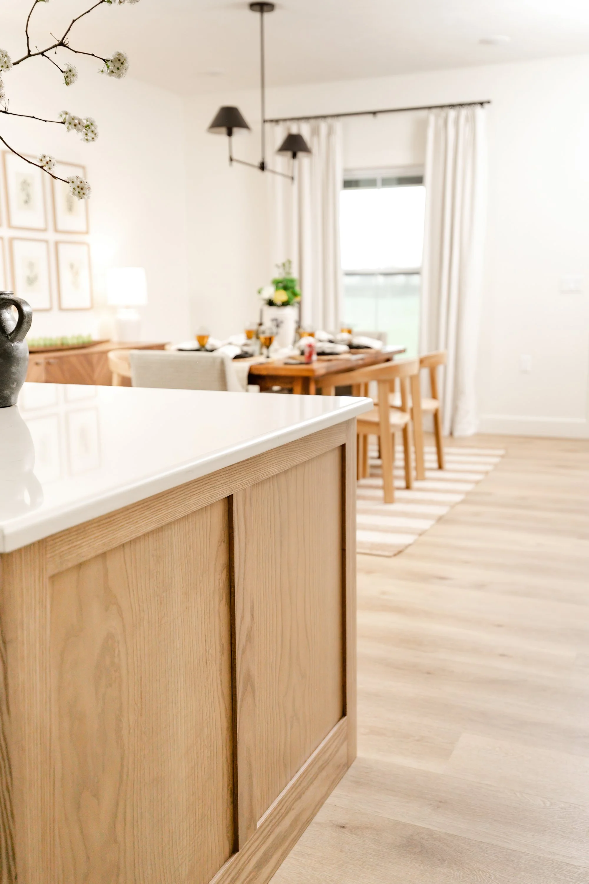 Close-up of a kitchen counter with a black pitcher, with a dining area in the background featuring a wooden table set for a meal, six glasses, and a plant in a white pot; white curtains cover a window in a bright, modern living space.
