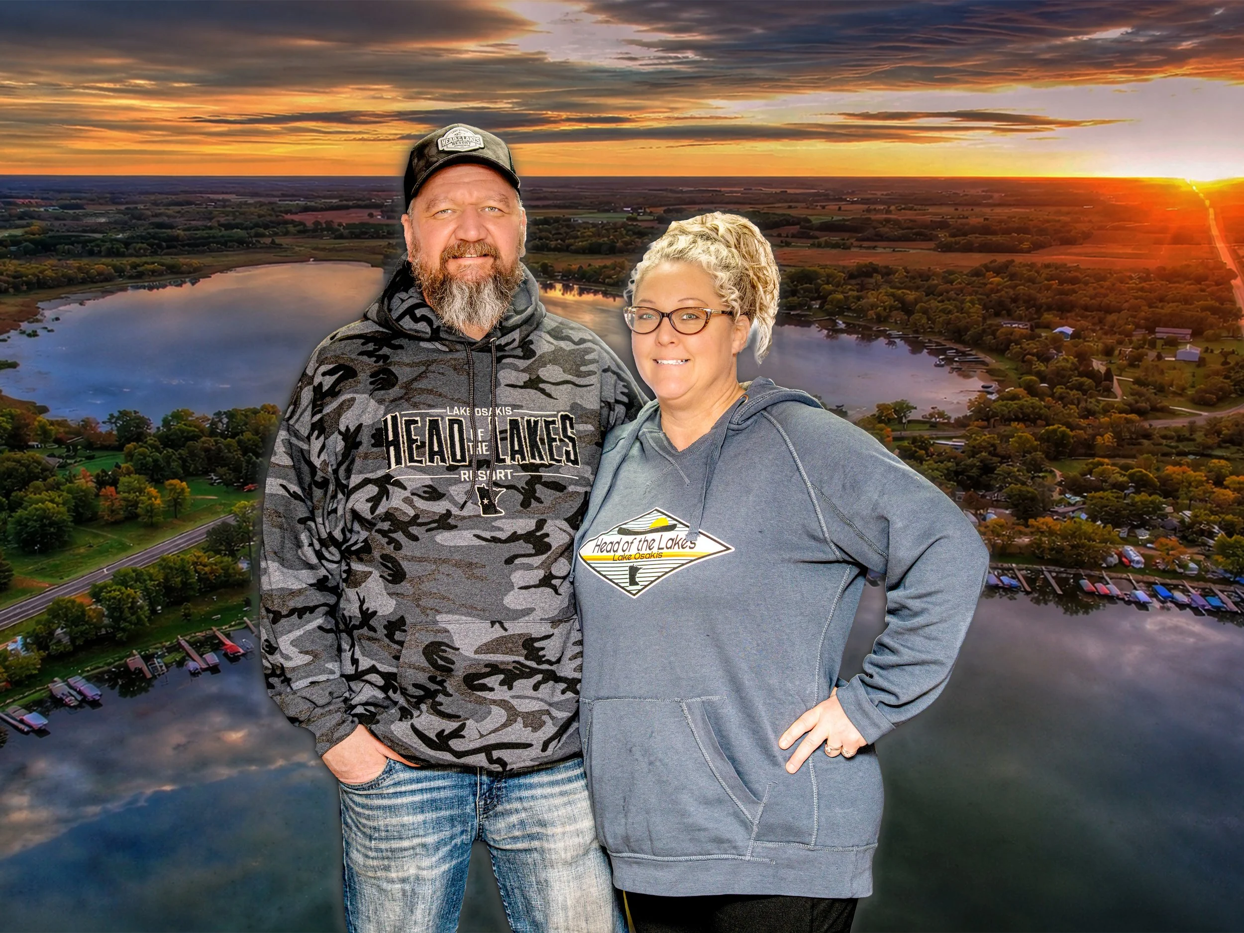 A man and a woman standing in front of an aerial view of a lake at sunset. The man is wearing a camouflage hoodie and cap, and the woman is wearing a gray hoodie with a logo that says 'Head of the Lakes Lake Osakis.'