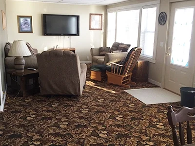 Living room with a floral carpet, a wall-mounted TV, two armchairs, a rocking chair, a side table with a lamp, and sliding glass doors leading outside.