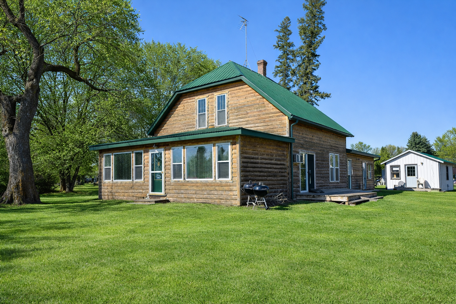 A two-story wooden house with a green metal roof, multiple windows with blinds, a small set of stairs leading to a door, and a grassy yard with trees and a small white building in the background.