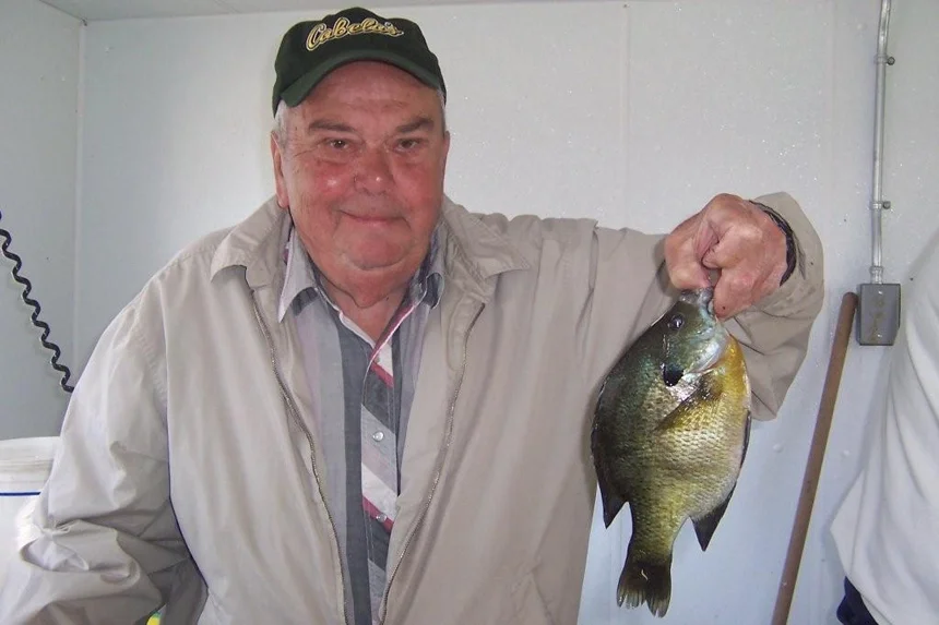 An elderly man wearing a Cabela's cap, smiling and holding a largemouth bass fish with his right hand.