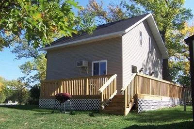 A two-story house with gray siding and a wooden deck in the backyard, surrounded by green grass and trees.