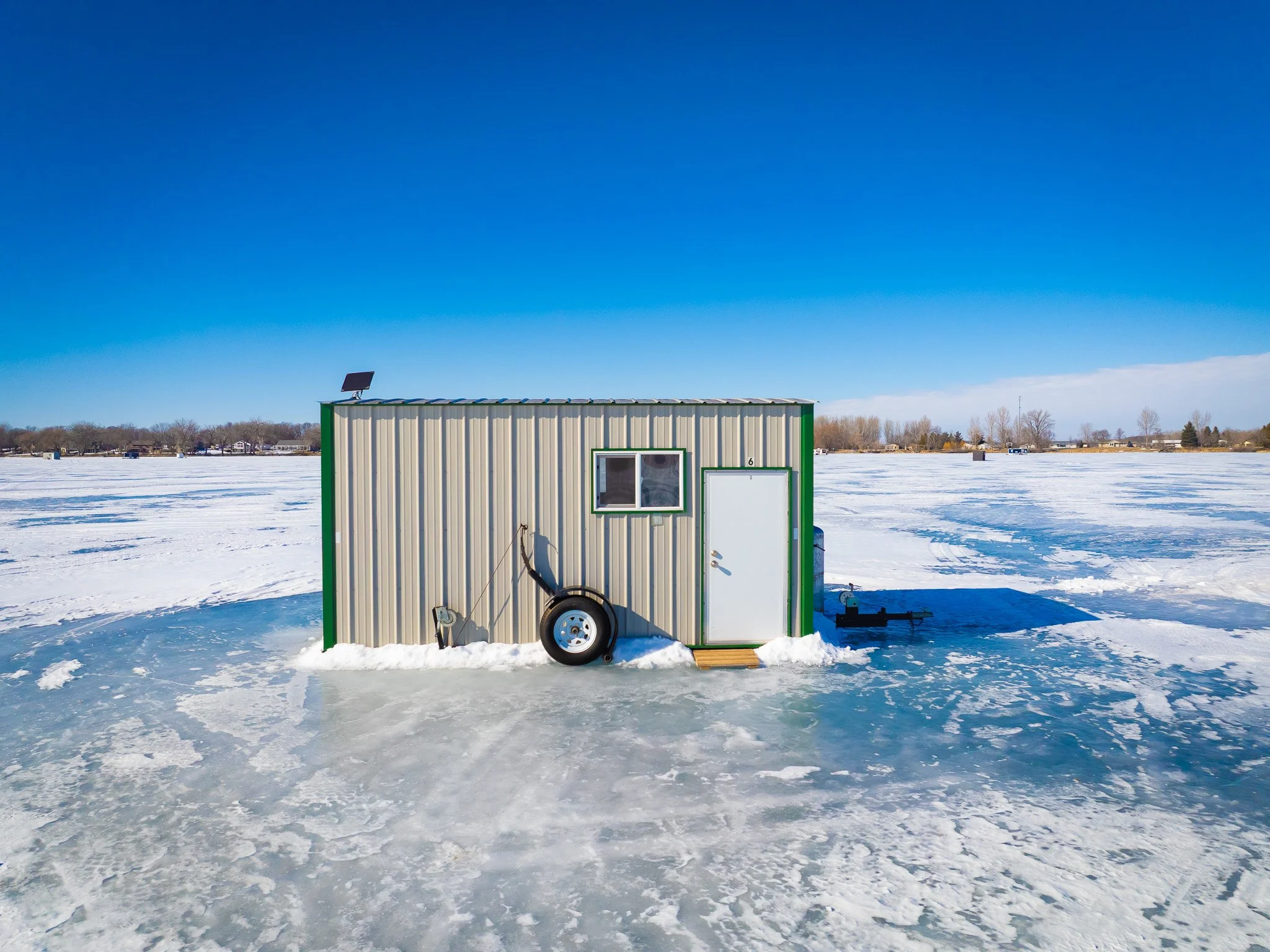 Small metal building on frozen lake with snow, clear blue sky, and distant trees and houses in the background.