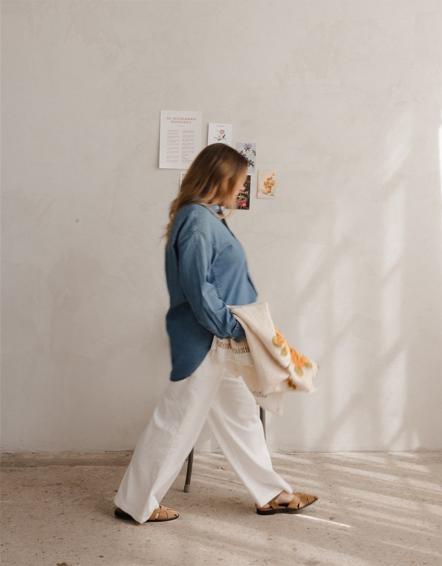 A woman walking indoors, holding a cloth bag, with a white wall in the background and a few printed papers or artwork taped to it.