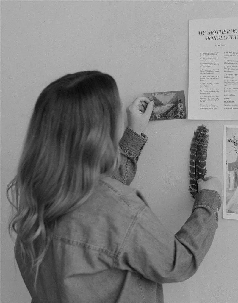 A person with long hair and a denim jacket is decorating a wall with various items including a postcard, a feather, and a printed poem titled "My Motherhood Monologue."