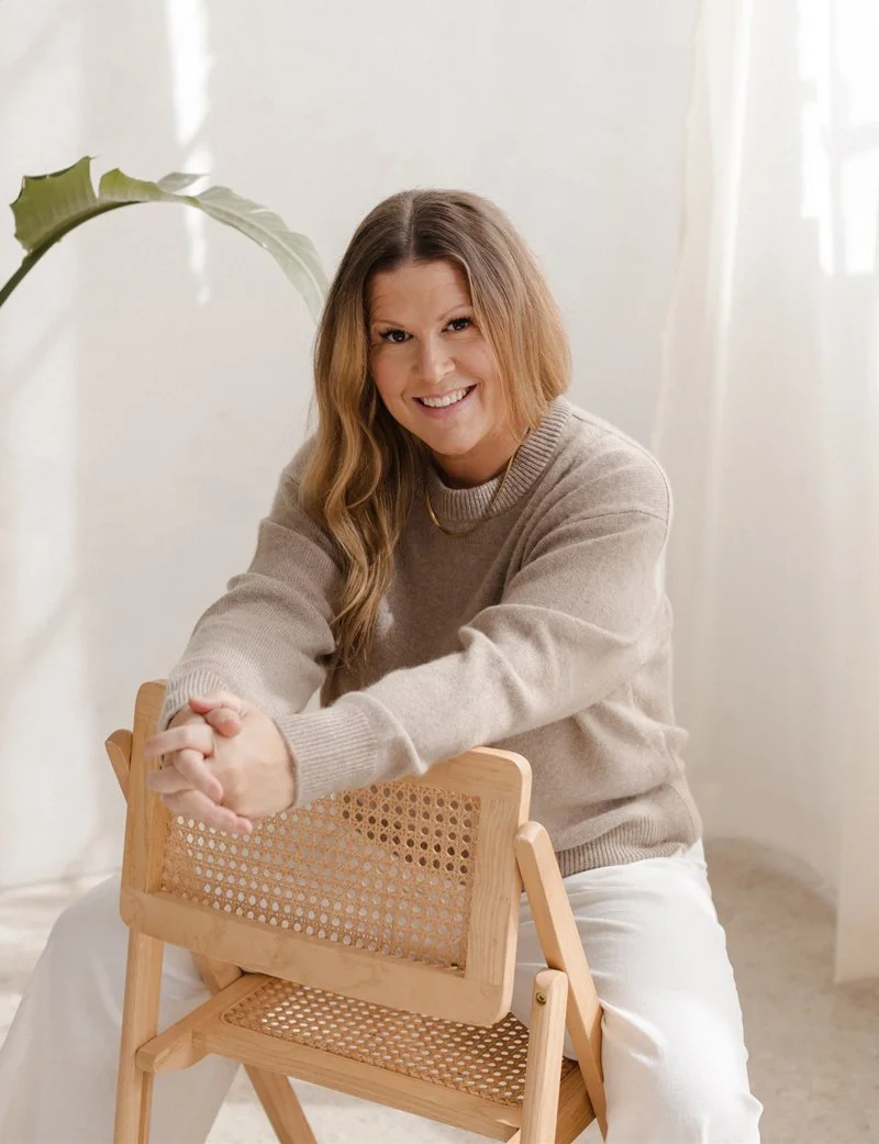 A woman with light brown, wavy hair sitting on a wooden chair with a woven cane backrest and seat, smiling and leaning forward with her hands clasped on the chair's back, in a bright, softly lit room.