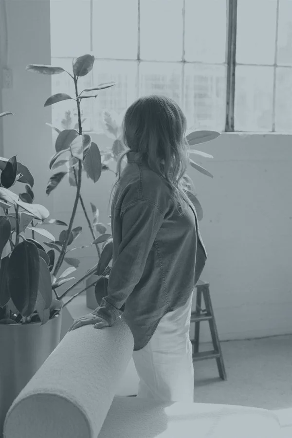 A woman with long hair, wearing a denim jacket and white pants, stands indoors near large windows and a potted plant, looking outside.