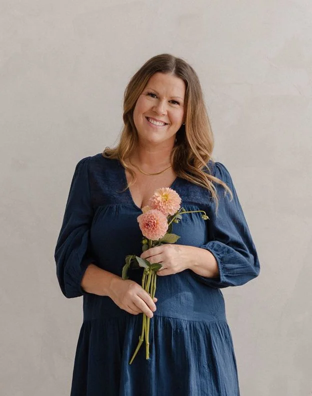 Smiling woman in a blue dress holding pink and peach flowers in front of a plain light-colored wall.