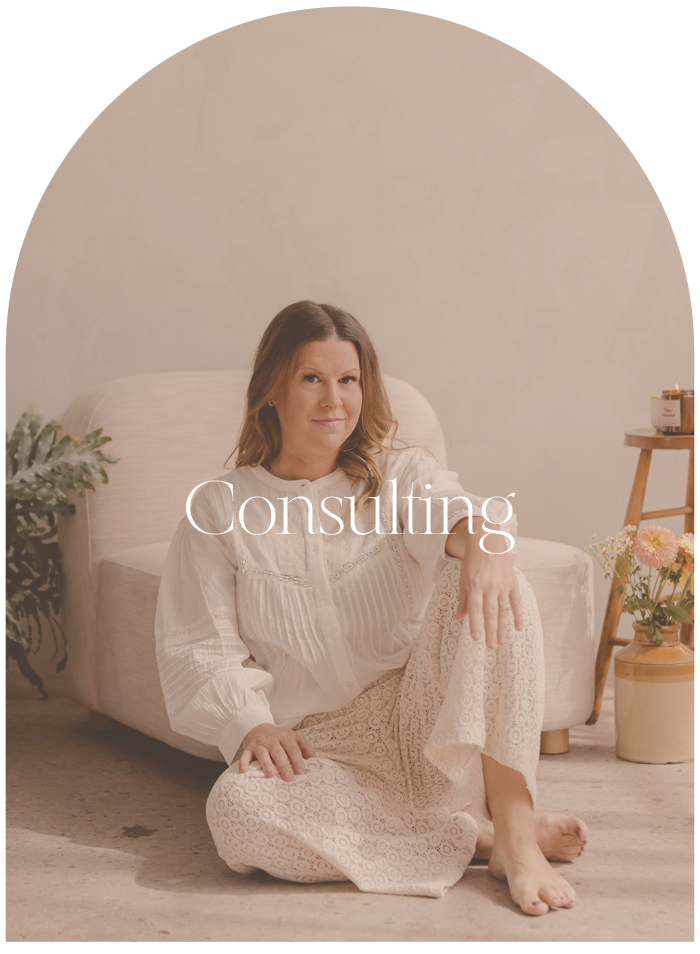 A woman sitting on the floor next to a beige sofa in a well-lit room, with a plant on her left and a small table with flowers on her right, with the word 'Consulting' overlayed.