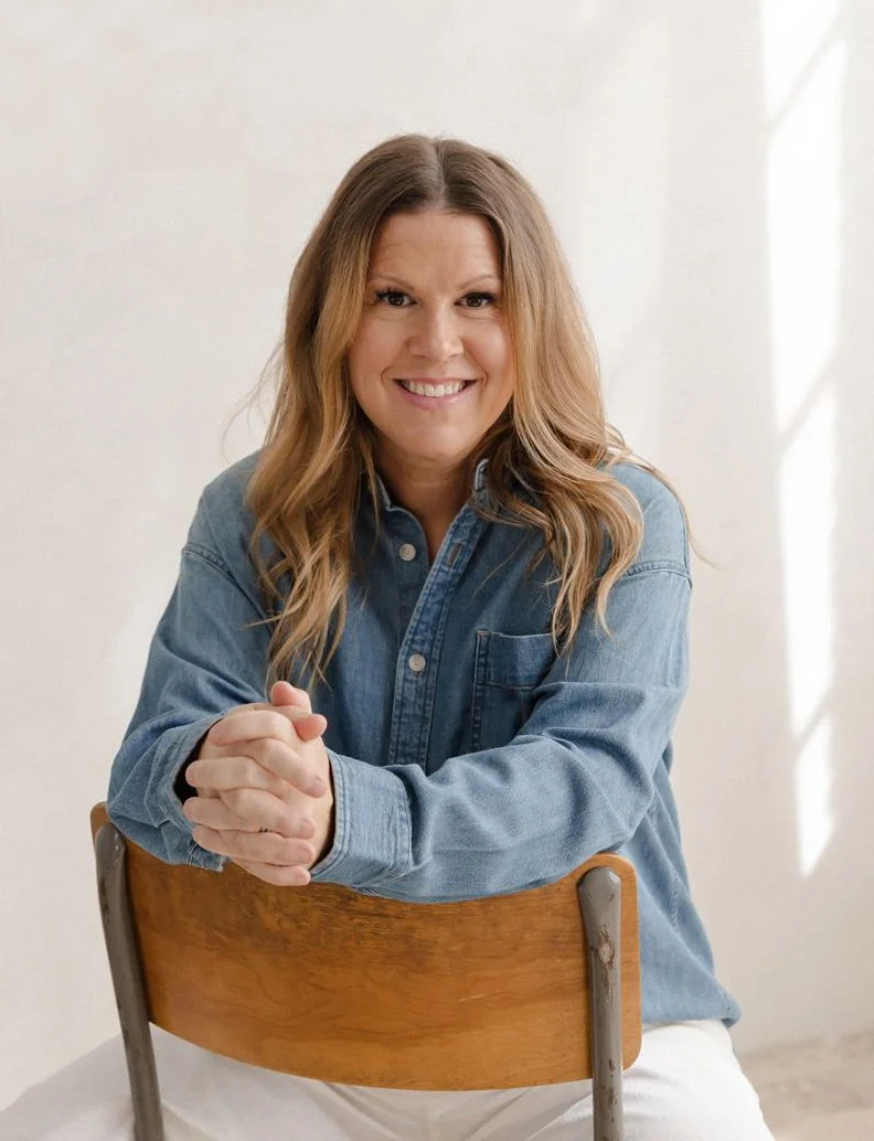 A woman with long, wavy brown hair, smiling and sitting on a wooden chair, wearing a blue denim shirt.