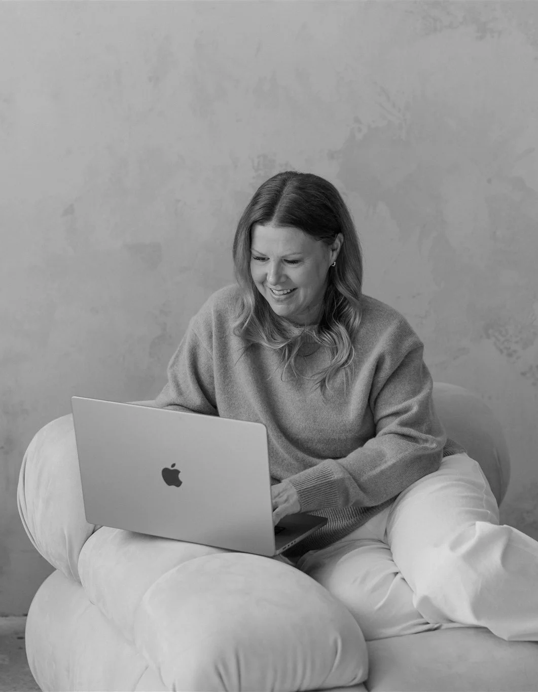 A woman with long, wavy hair sitting on a plush cushion or bean bag, working on a MacBook laptop, smiling, against a plain wall.