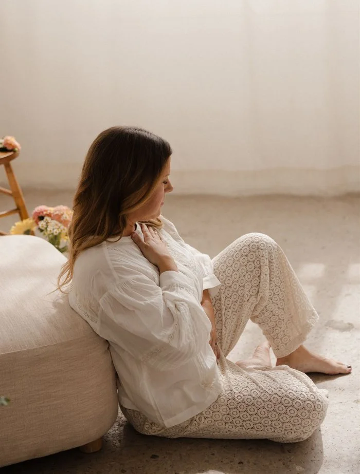 A woman sitting on the floor leaning against a large beige cushion, wearing white pajamas with a delicate lace pattern. She has long, wavy brown hair and appears to be in a peaceful, reflective mood. There are flowers in the background.