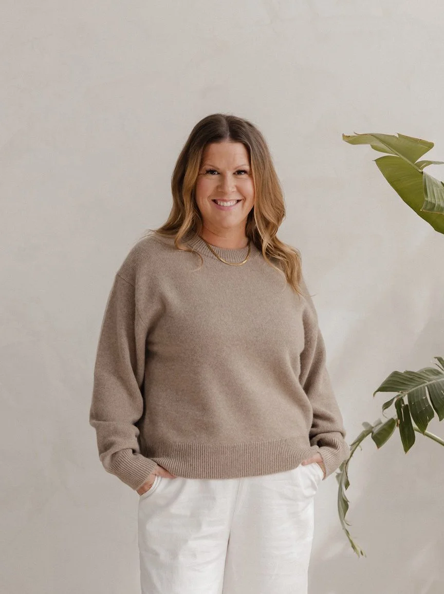 A smiling woman with shoulder-length wavy brown hair stands against a neutral background, wearing a beige sweater and white pants, with a gold necklace. There is a green potted plant to her right.