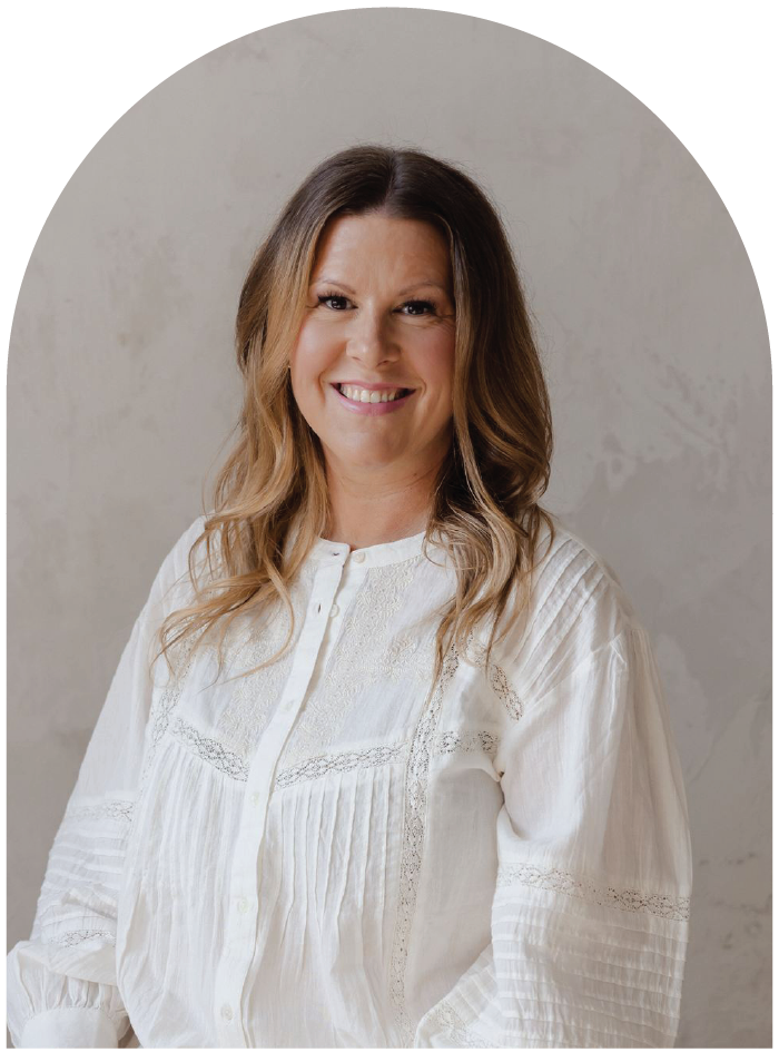 A woman with long, wavy blonde hair smiling, wearing a white button-up blouse with lace and embroidery details, against a light, textured background.