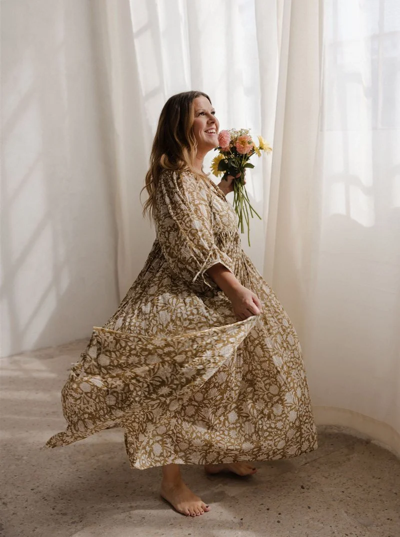 Woman in a floral patterned dress holding a bouquet of flowers and smiling near white curtains.