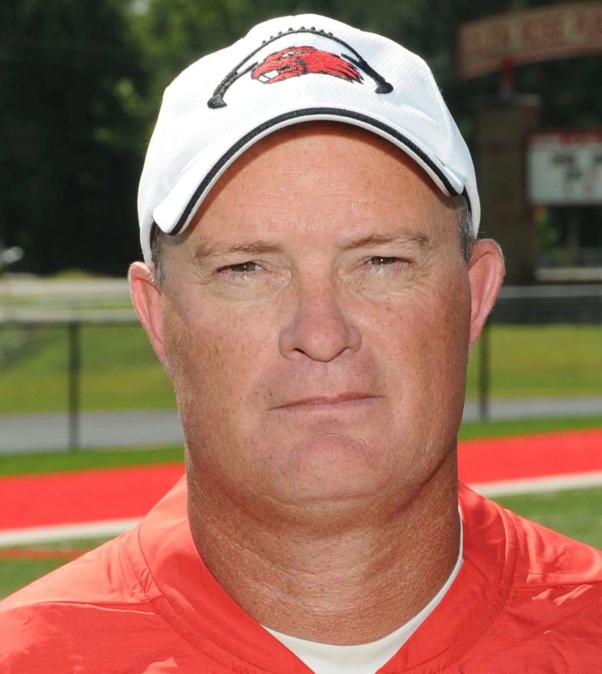 A man wearing a white baseball cap with a red and black logo and an orange sports shirt, standing outdoors on a sports field.