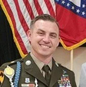 A man in a military uniform smiling, standing in front of American flags.