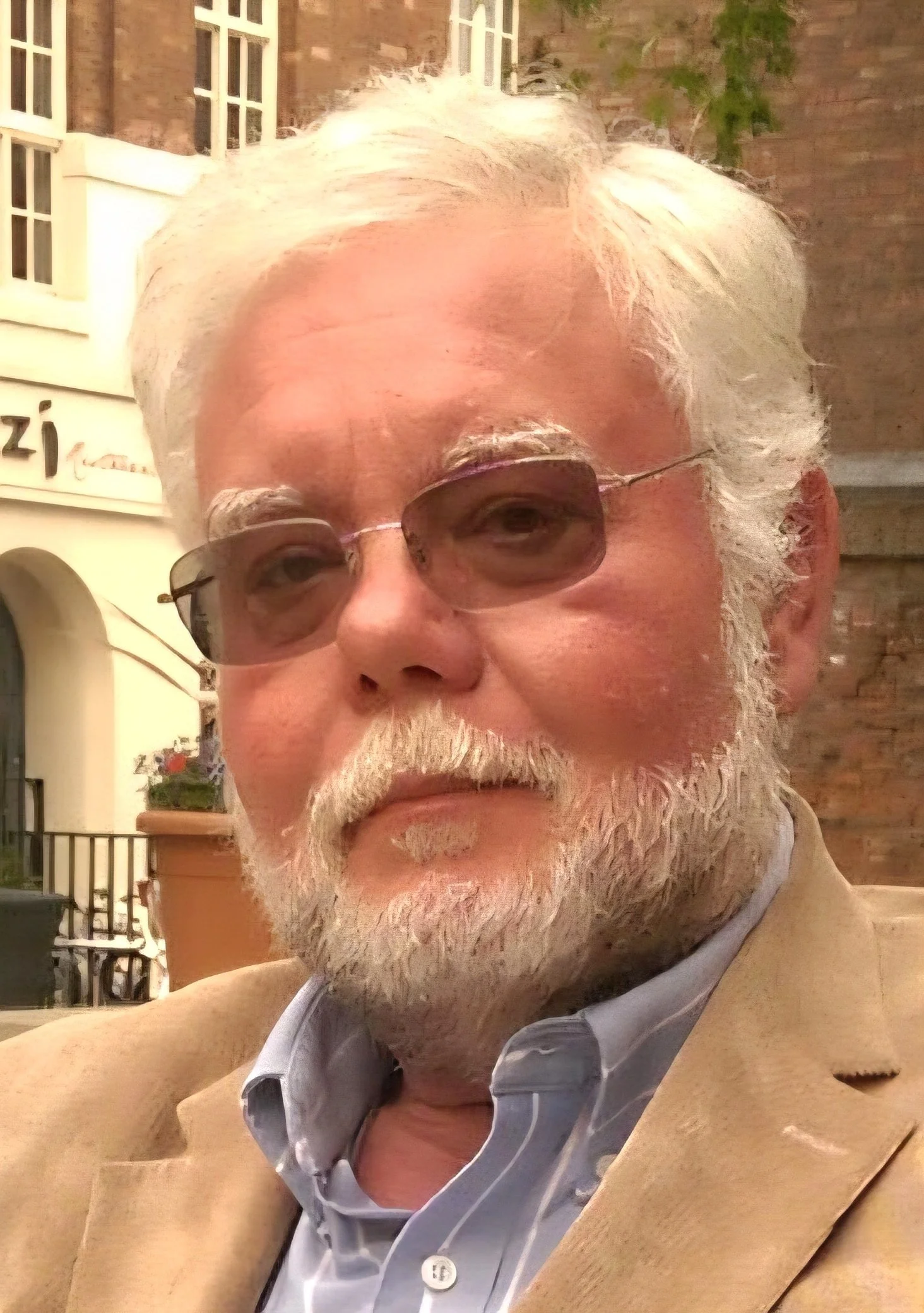 Close-up of an elderly man with white hair and beard wearing sunglasses, a beige blazer, and a light blue collared shirt, outdoors in front of a building and some greenery.