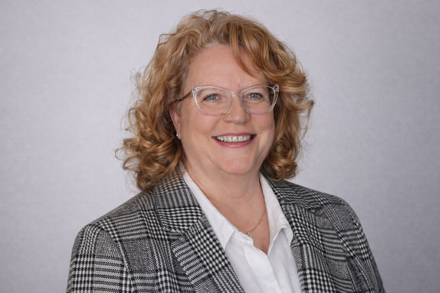 A smiling woman with curly red hair, wearing glasses, a white shirt, and a plaid blazer, standing in front of a plain light gray background.