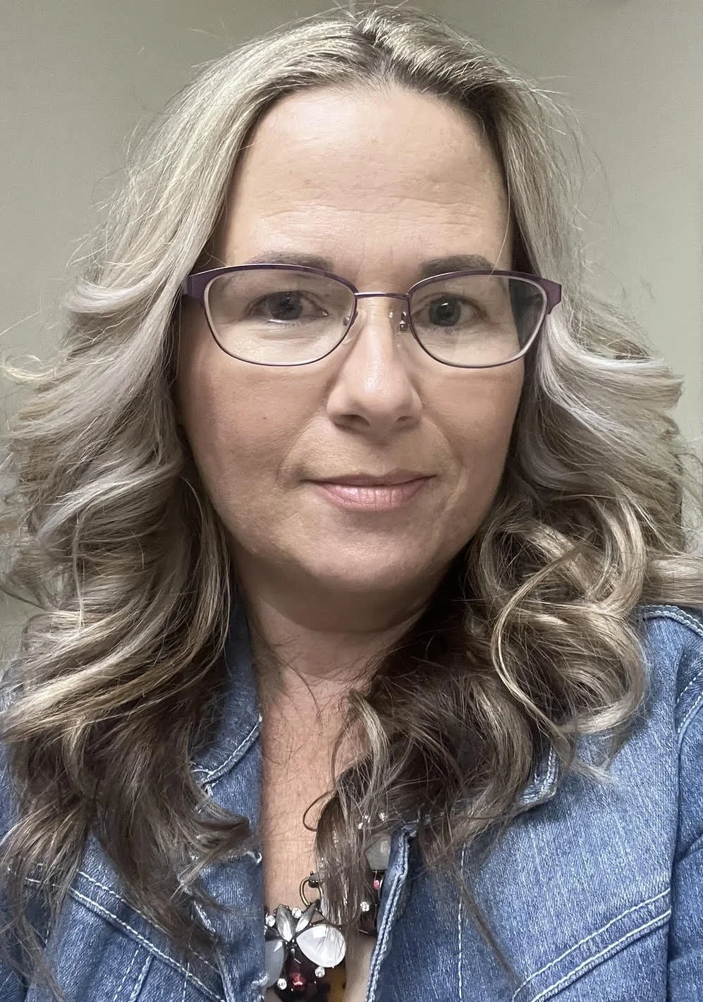 A woman with wavy, shoulder-length blonde hair and glasses, wearing a denim jacket and a necklace, smiling at the camera.