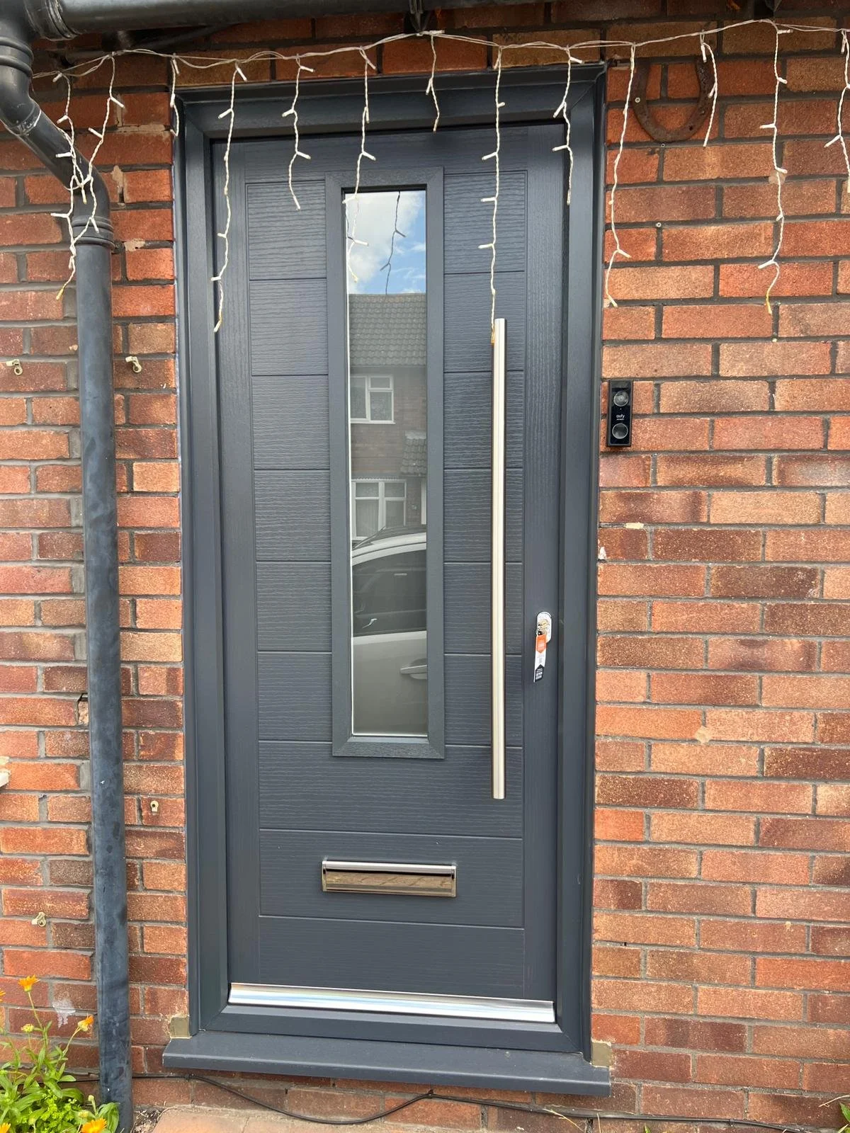 Modern front door with a vertical glass window and silver handle, set in a brick wall, decorated with string lights.