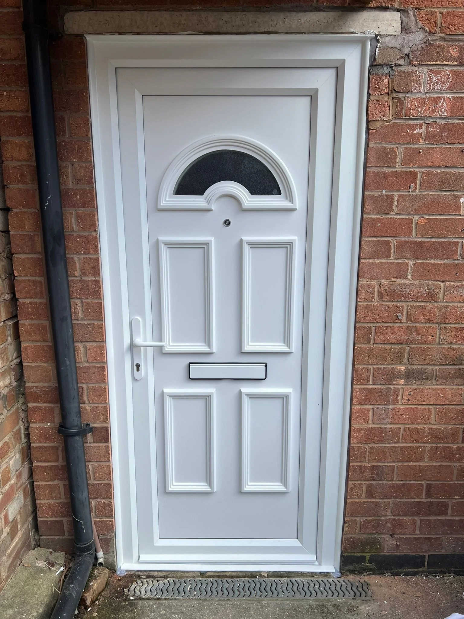 White front door with decorative rectangular panels, a semi-circular window at the top, a mailbox slot, and a door handle, set in a brick wall with a downspout on the left.