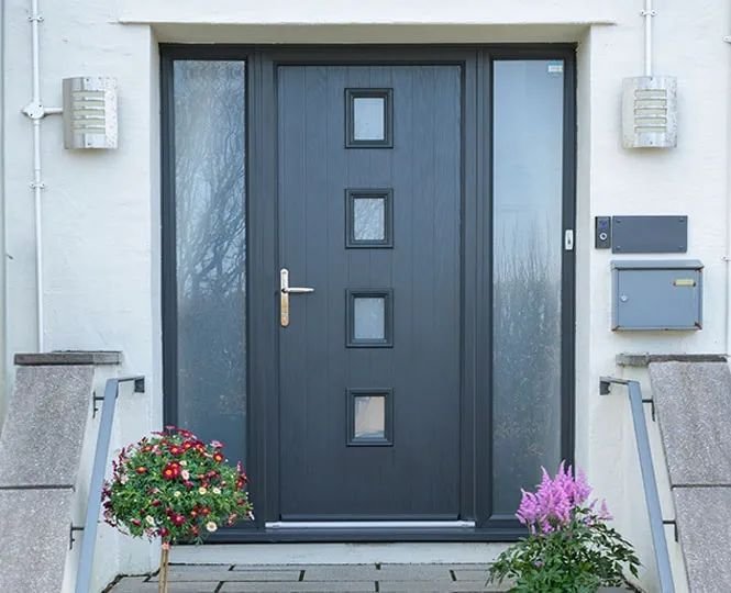 Black front door with four small square windows, flanked by two outdoor wall lights, with a mailbox and doorbell to the right, pink and red flowers on either side, and concrete steps leading up to the door.