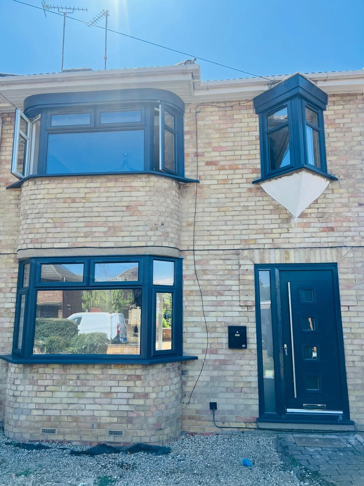 The front of a brick house with black window frames, a black front door with square windows, and two bay windows, one on the ground floor and a smaller one above on the second floor. The house has light-colored bricks and there is a power line and an