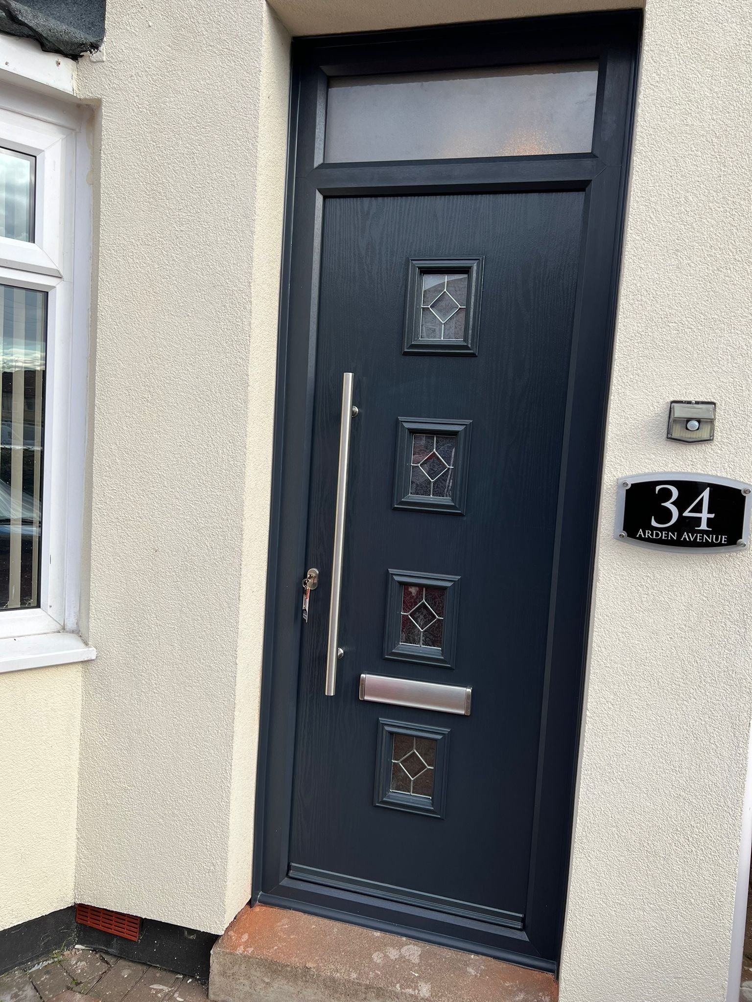Black front door with three decorative glass panels, a vertical handle, a mail slot, and a set of keys hanging from the handle. The door is outside on a beige wall with a house number plaque displaying "34 Arden Avenue."