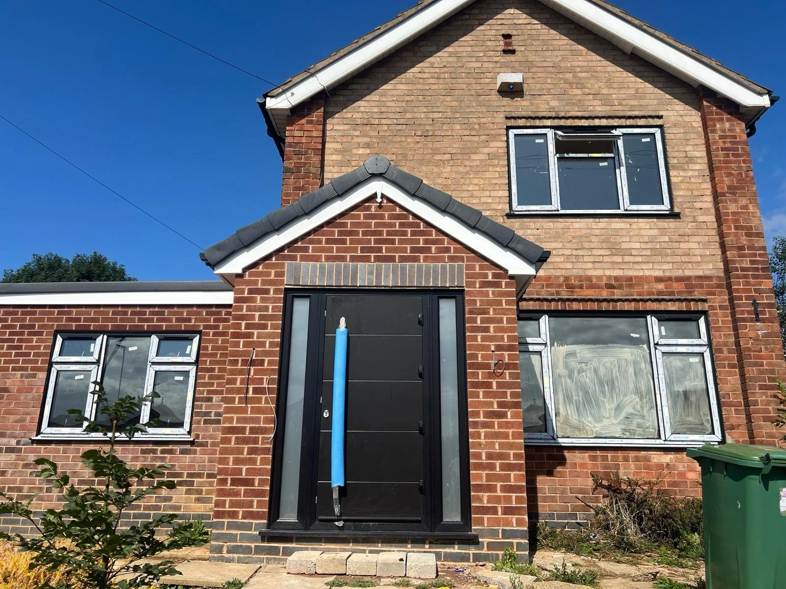 A two-story brick house under construction with a black front door, three windows with open panels, and a green trash bin nearby, set against a clear blue sky.