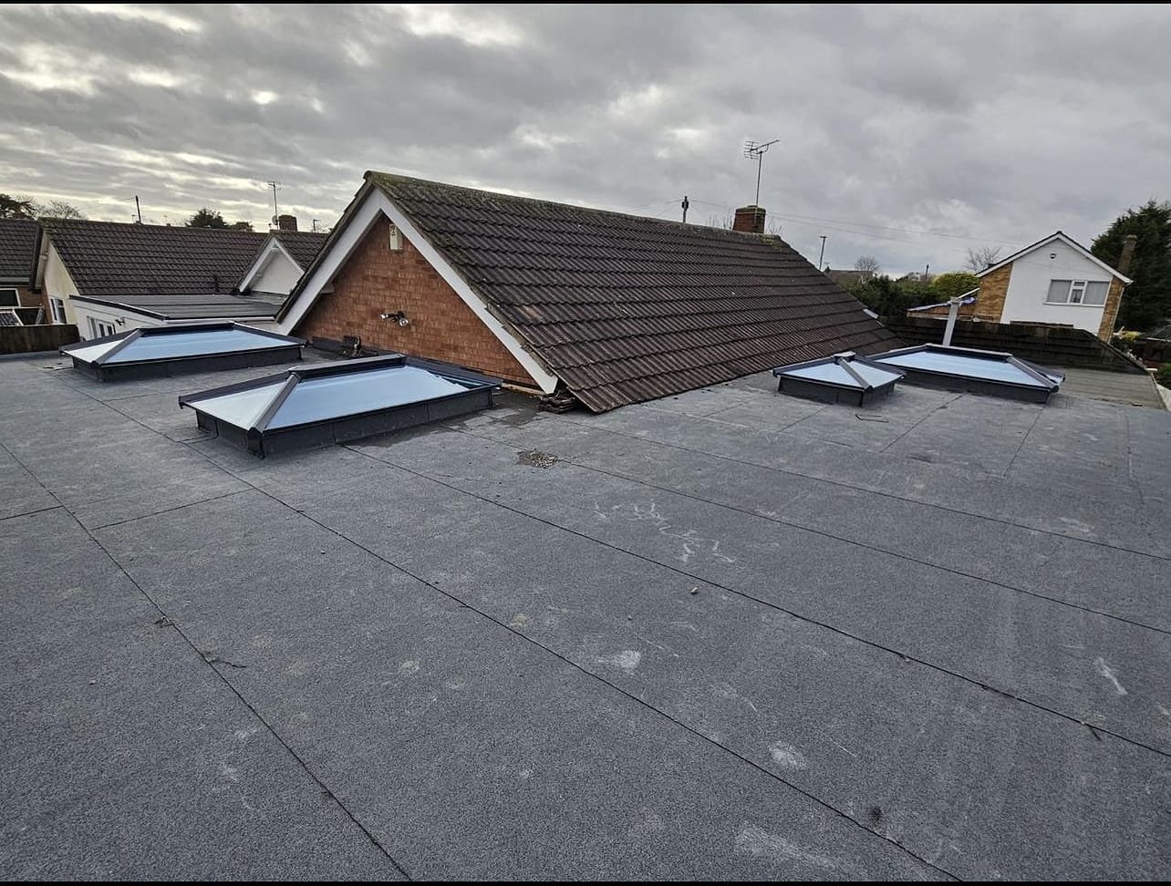 View of a flat roof with four skylights and a sloped brick house roof in the background, under cloudy sky.