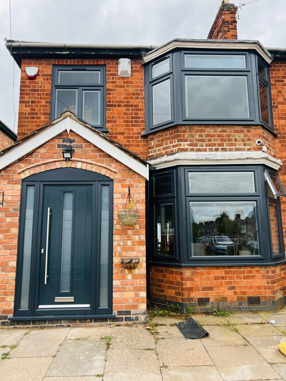 Front of a brick house with blue door and bay windows, gray roof, hanging plant, and small waste bin on sidewalk.