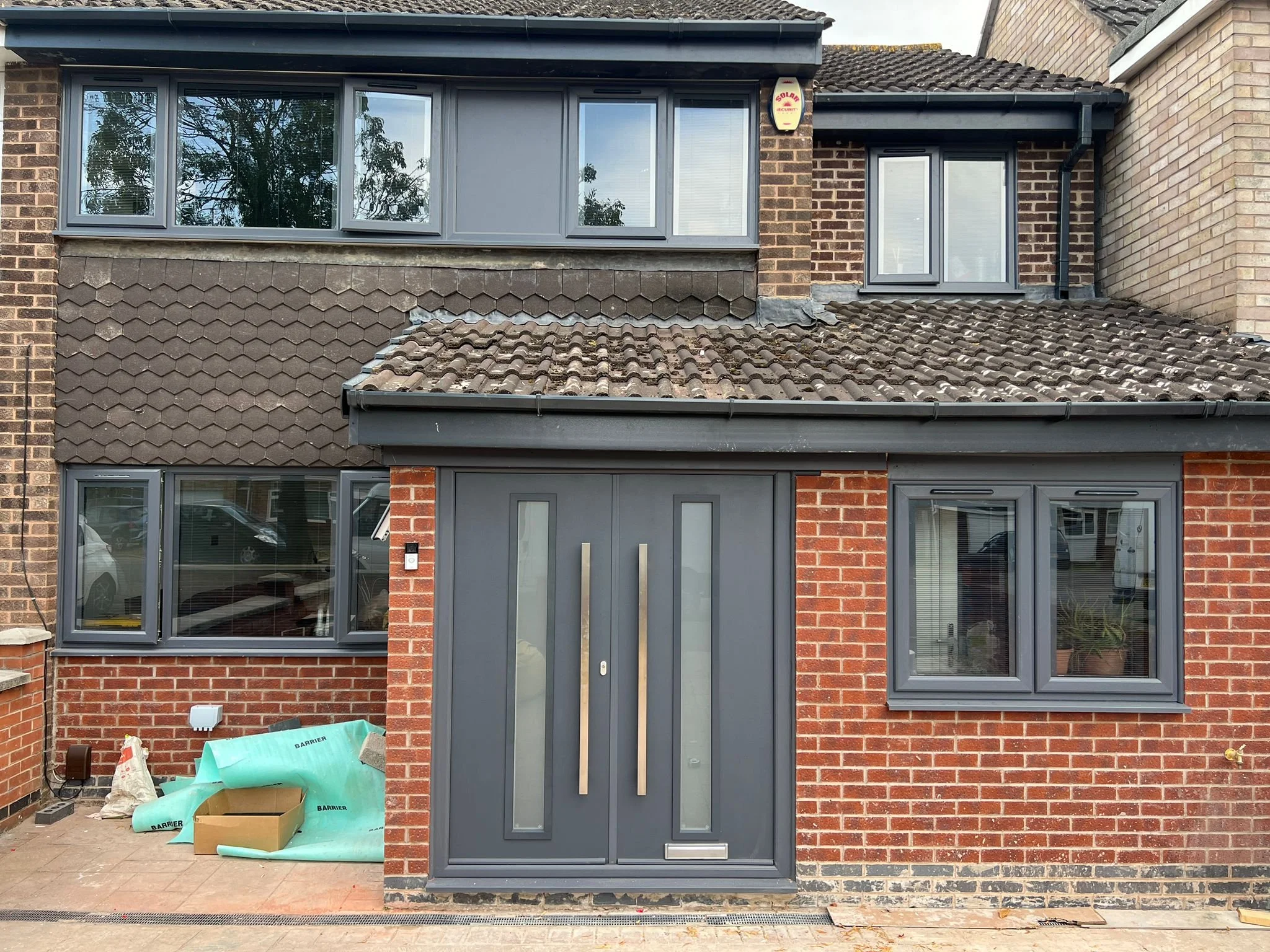 Front view of a two-story brick house with gray window frames and a gray front door. Windows on both floors, some open, and construction materials on the ground near the house.