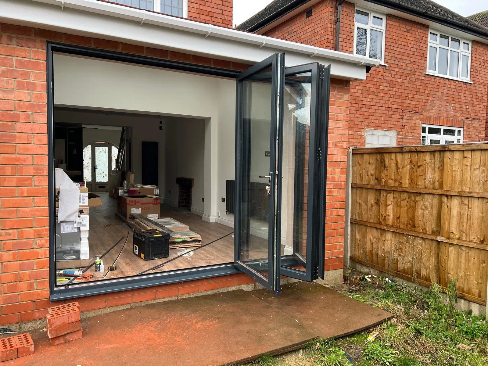 The back of a red brick house with a large, open double-glazed glass door leading to an interior room under renovation, with construction tools and materials inside. There is a wooden fence on the right and a small patch of grass and dirt outside.