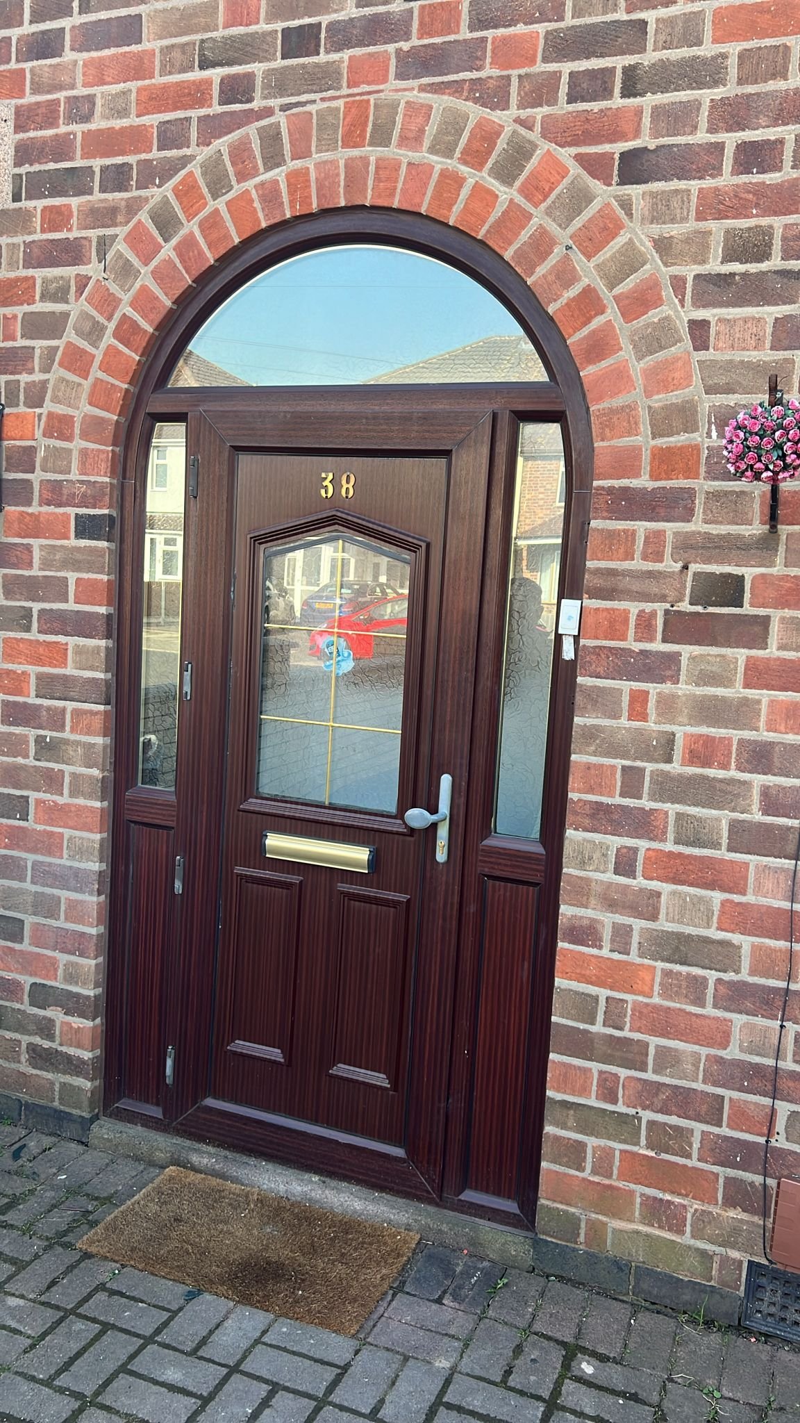 Front door of a house with a brick exterior and an arched window above, featuring a wooden door with the number 38, a mailbox slot, a handle, and a frosted glass panel, with a small flower arrangement on the side.