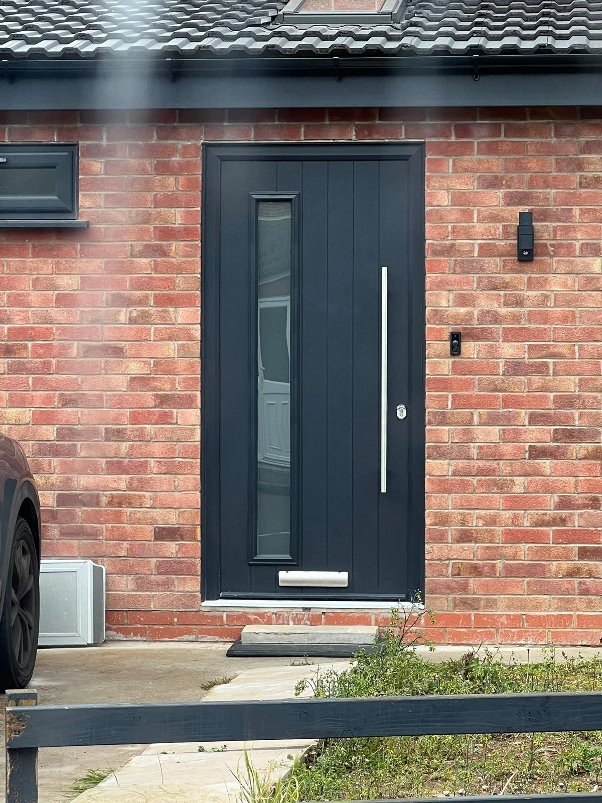 A black front door with a vertical handle and a narrow window on a brick house exterior, with a small garden in front.