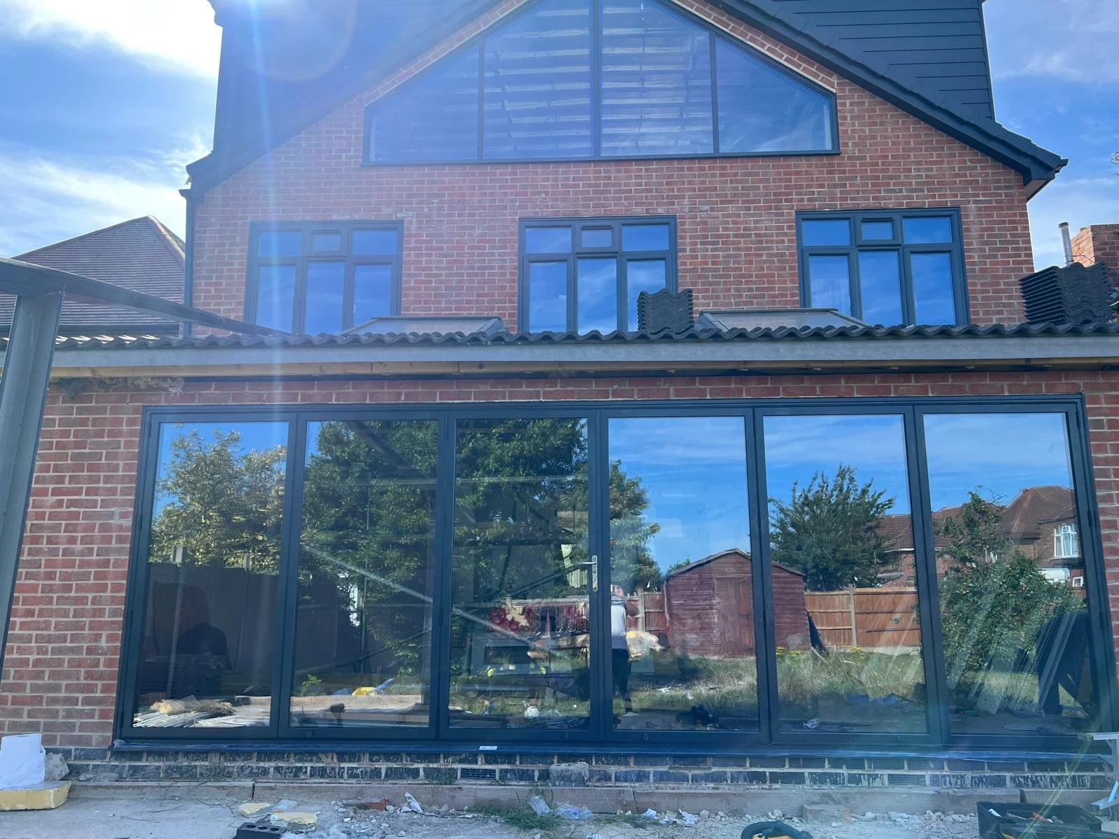 Construction site of a brick house with large glass windows and doors, with a garden shed and trees visible in the background under a blue sky.