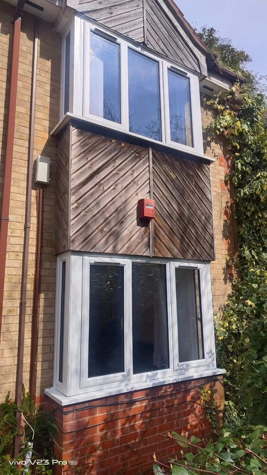 The image shows the exterior of a brick house with two white-framed bay windows, one on the lower part and one on the upper part, and a wooden panel section in the middle with a red alarm box attached to it. There are green plants and ivy on the side