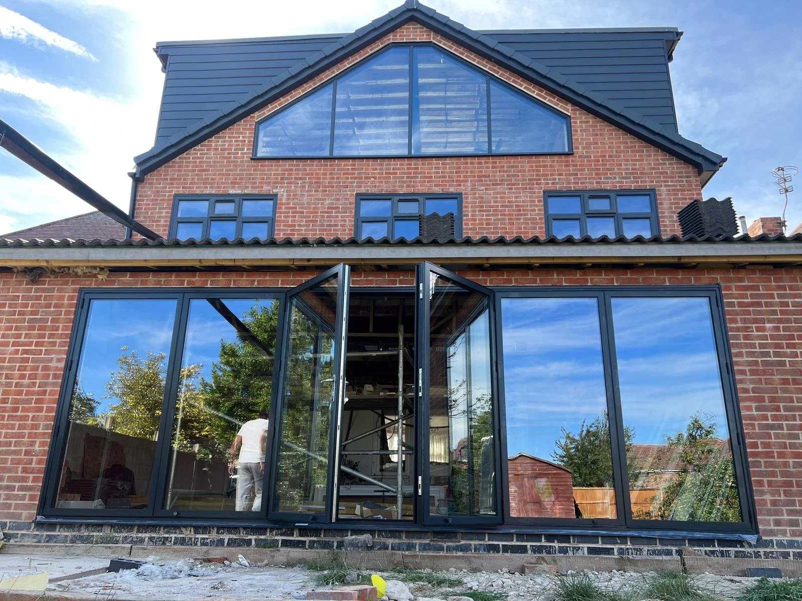 The image shows a multi-story brick house with black-framed glass windows and doors. The house has a brick exterior with a black roof. The front of the house has large glass doors, with one door open, revealing an interior space under construction or
