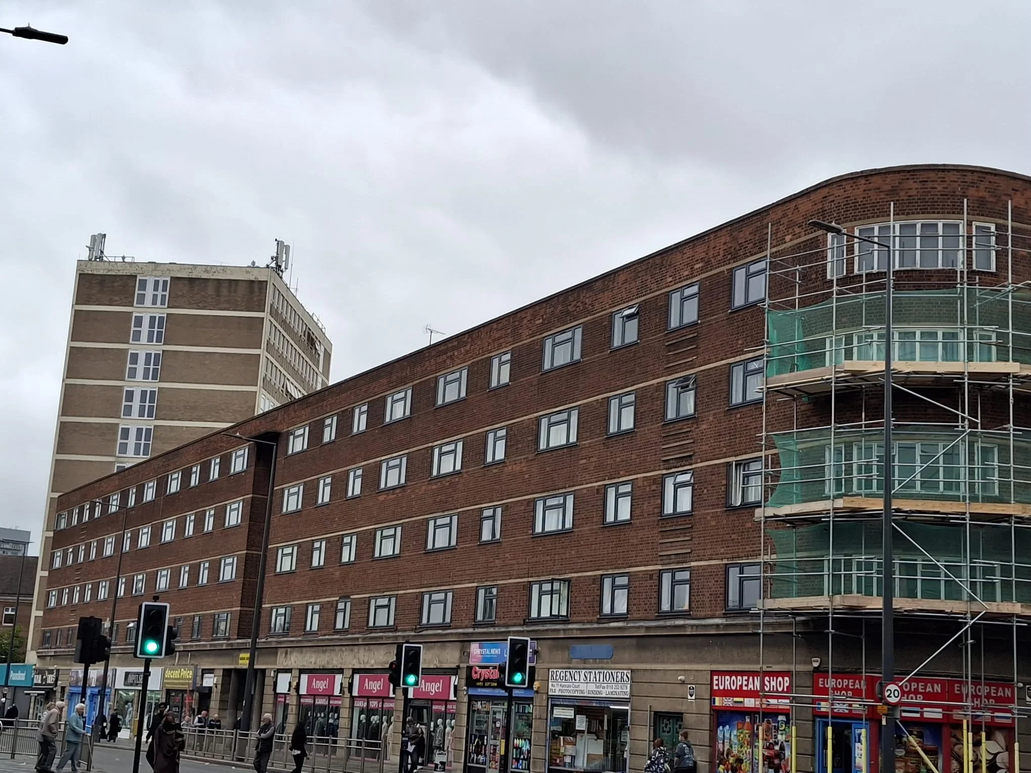 City street scene with a multi-story brick building under construction, scaffolding on the right, and shopfronts at the ground level. Pedestrians are walking on the sidewalk, and traffic lights are visible.
