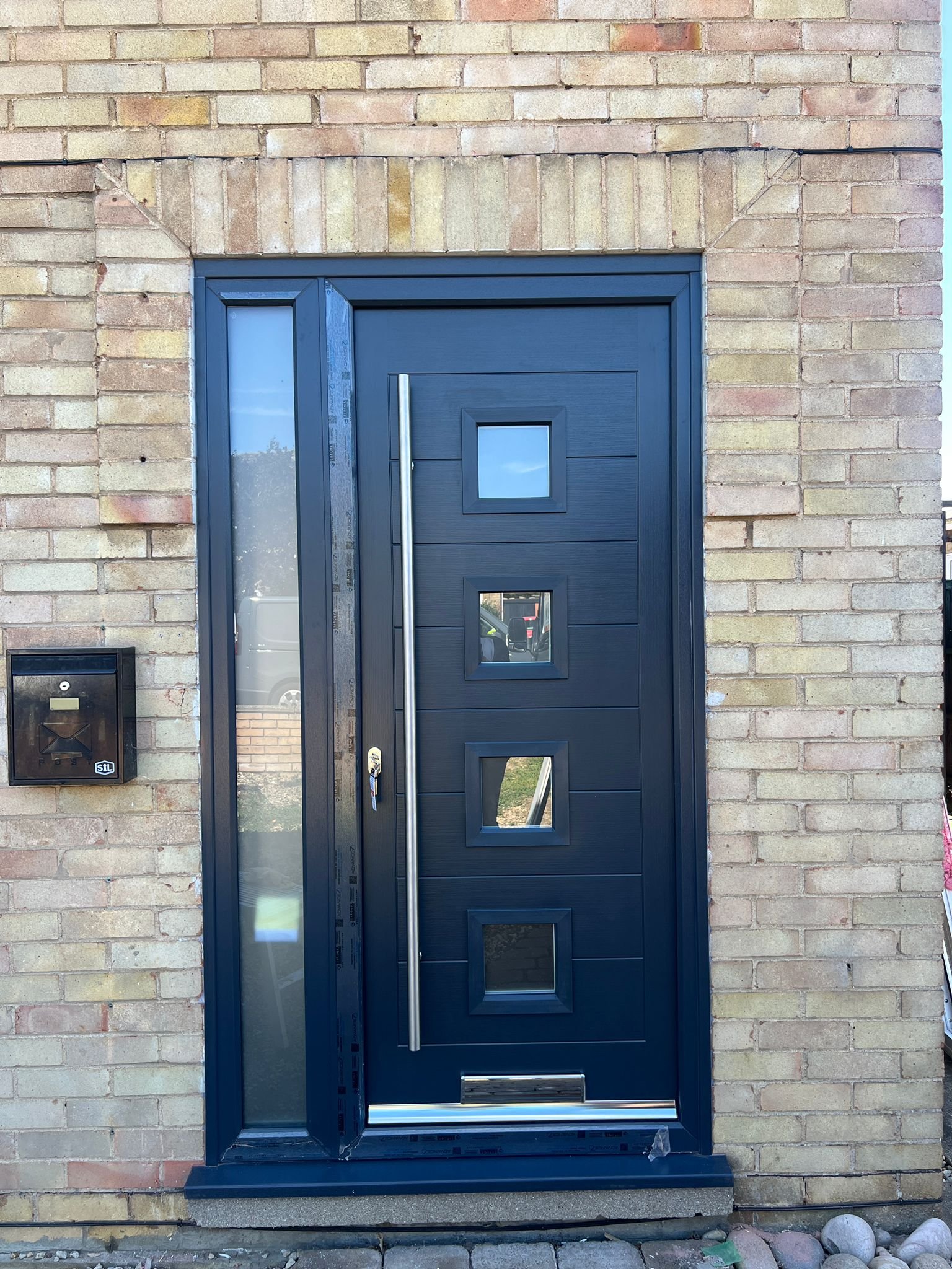 Modern navy blue front door with four small square windows, vertical handle, and mail slot, set in a brick wall with a mailbox to the left.