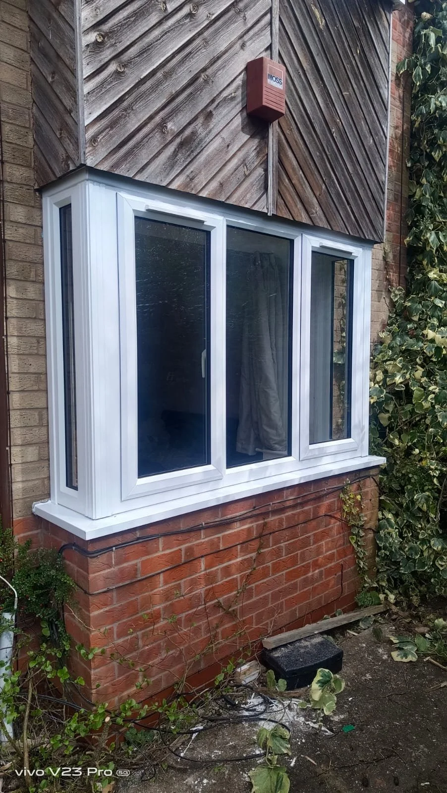 A brick house exterior with a white sliding window and a wooden panel section above. There is a small red device mounted on the wall above the window, and some overgrown ivy on the right side. The ground has some dirt, leaves, and electrical wires.