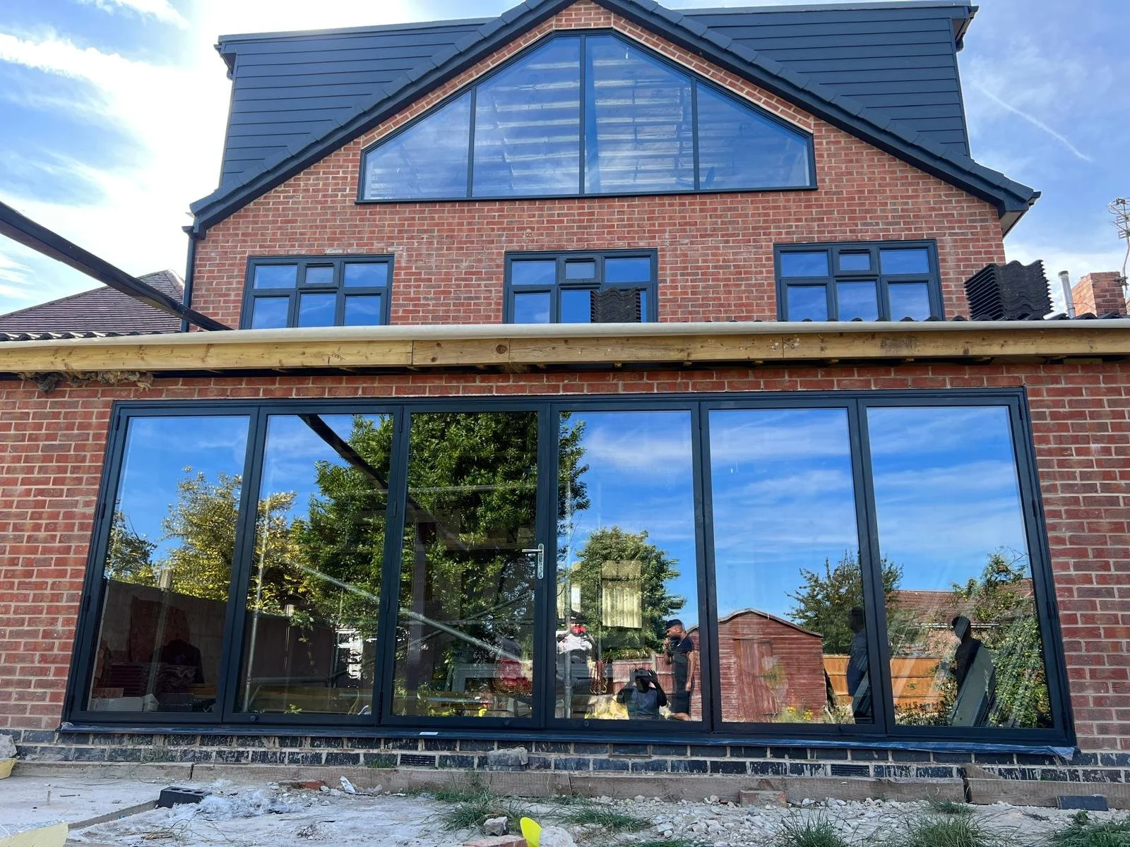 A brick house with large windows, including a prominent upper triangular window, with a newly installed glass extension on the ground level reflecting trees and the sky. Construction work is ongoing in the backyard.
