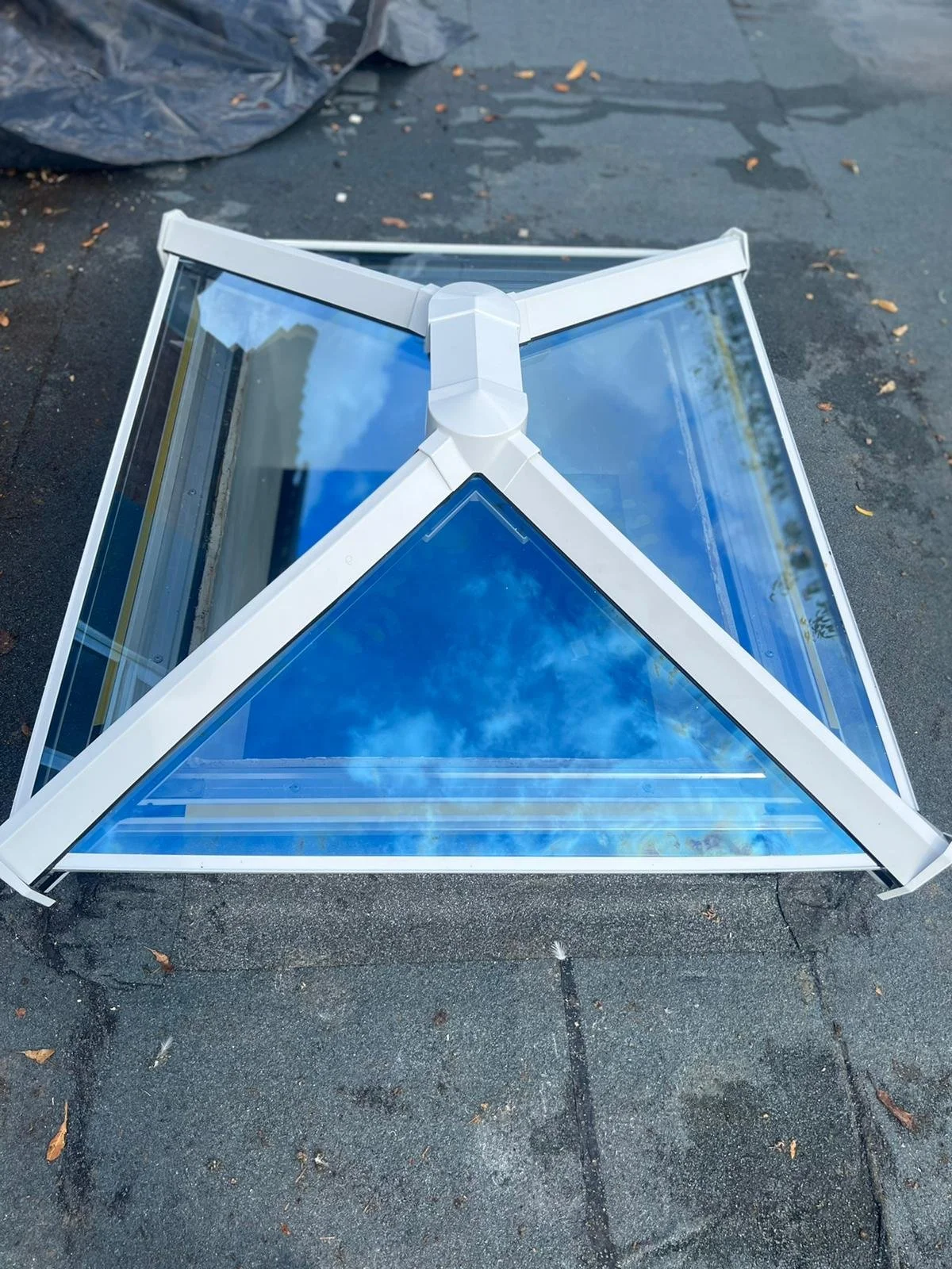 A white-framed glass skylight window with multiple panes reflecting a blue sky with clouds, placed on a paved surface outdoors.