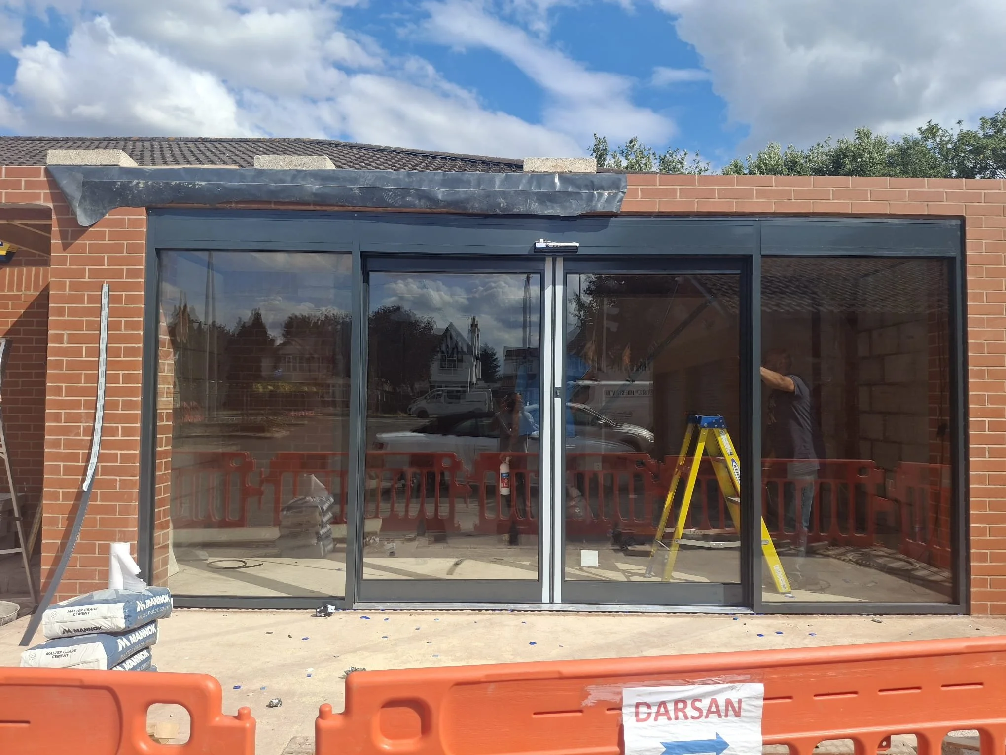 Construction site with large glass sliding doors being installed on a brick building. Construction materials and tools are visible inside and outside, with safety barriers and a ladder present.