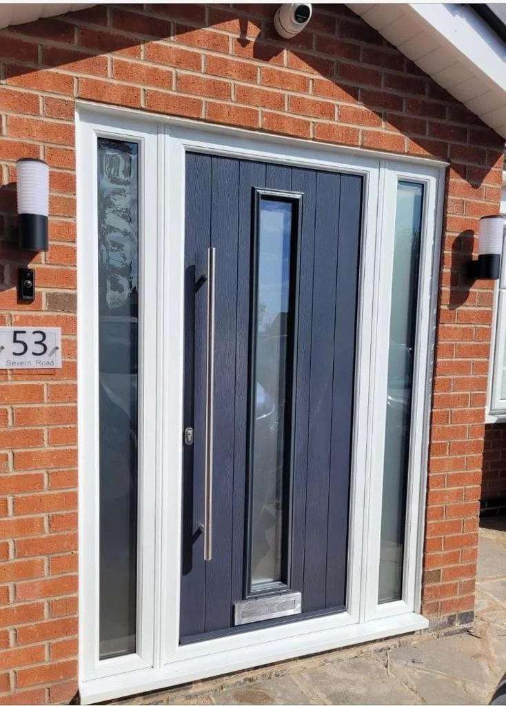 Modern front door with dark blue panel, vertical window, silver mailbox, and black handle, surrounded by a white frame, on a brick wall with two wall-mounted lights.