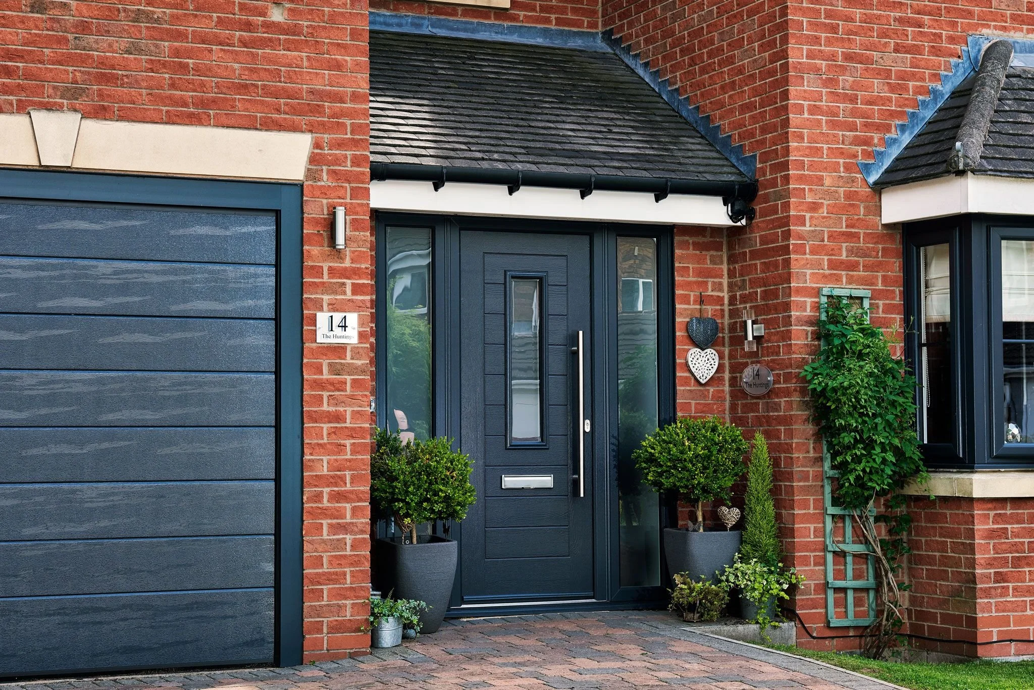 A modern brick house with a black front door, surrounded by potted plants and decorative hearts on the wall.