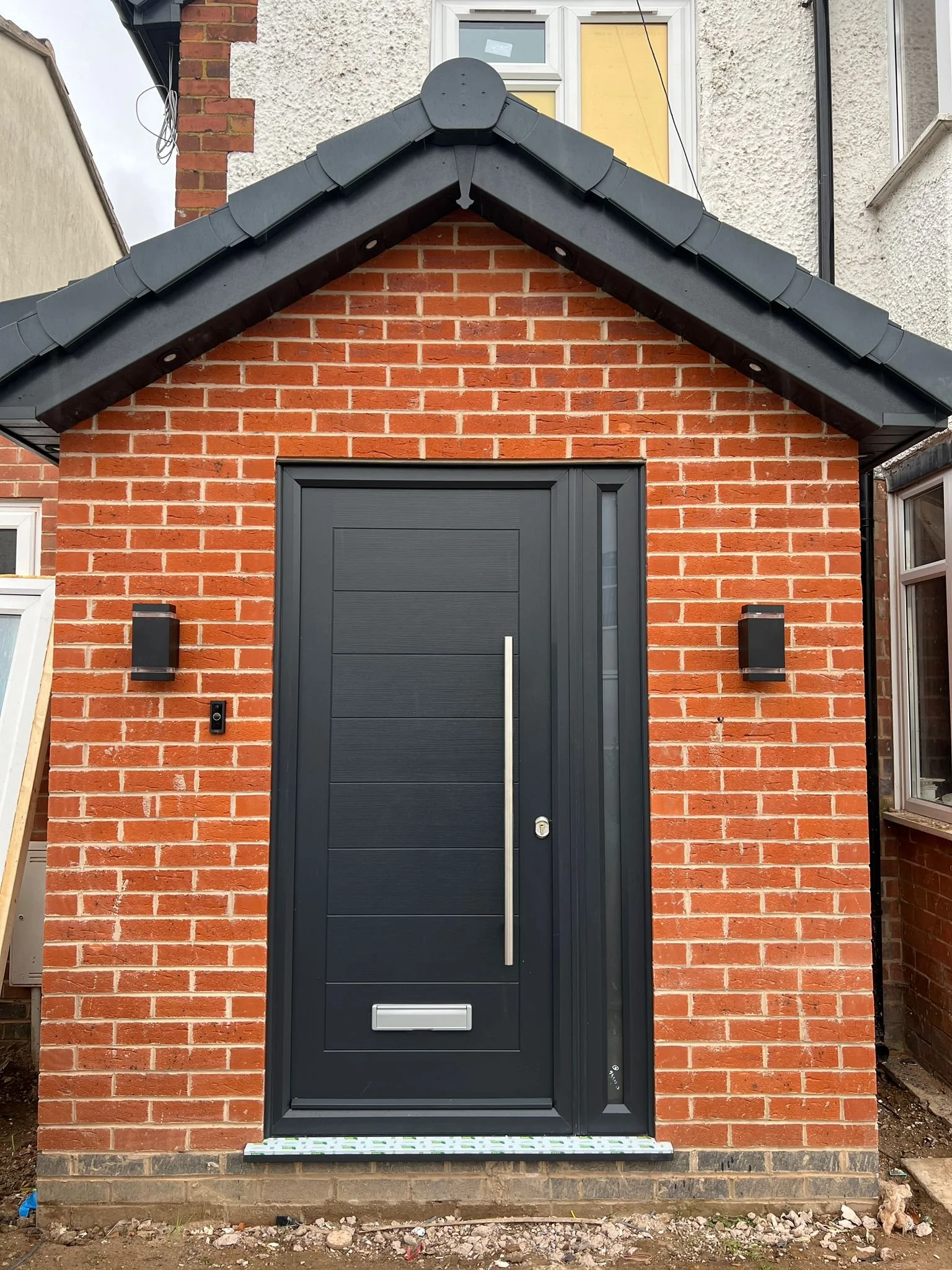 Front view of a small brick building with a black door, two black wall-mounted lights, and black roof tiles. An address bell and mail slot are also visible.