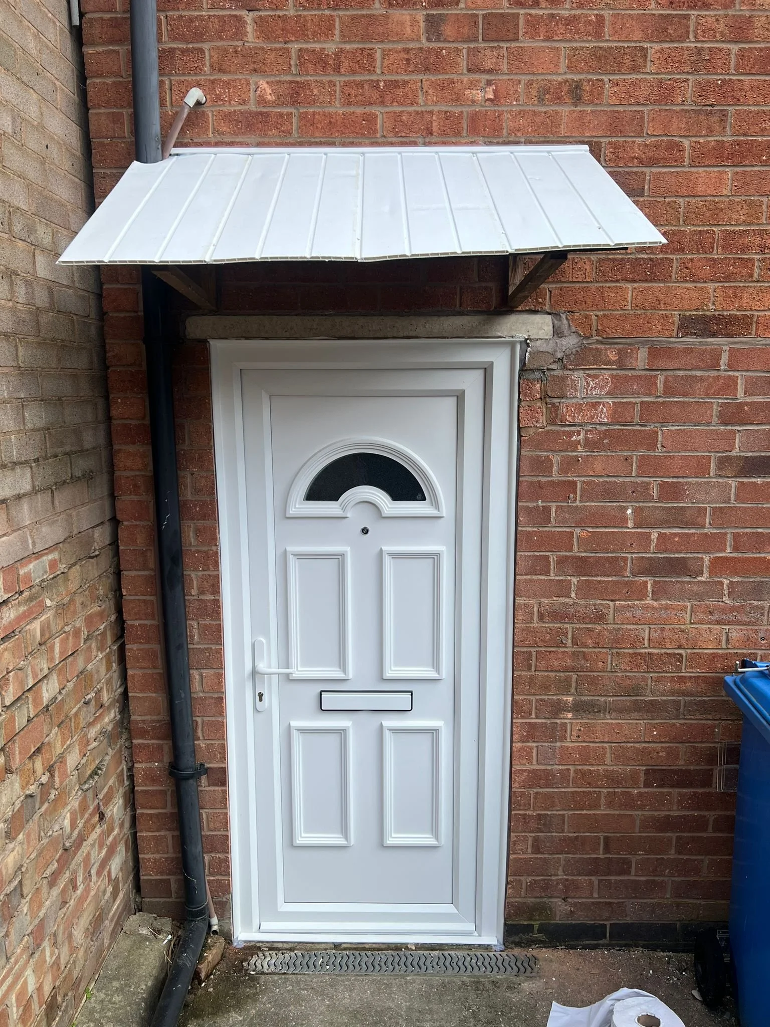 White door with decorative panels and a semi-circular window at the top, set in a brick wall, with a small overhang roof above.