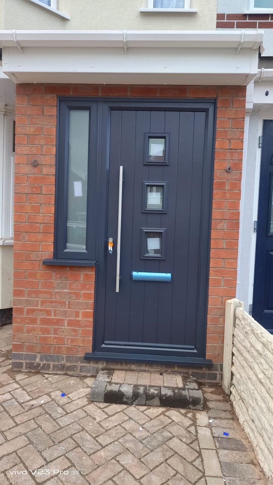 Black front door with three small square windows, a vertical silver handle, and a blue mail slot, set in a red brick wall with a window to the left and a white overhang above.